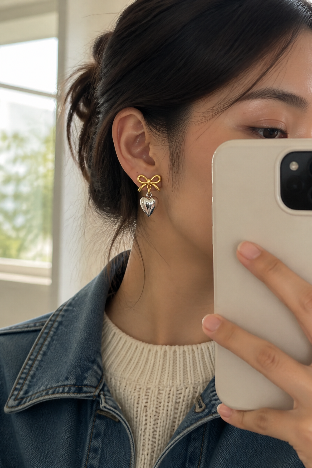 Southeast Asian woman taking a mirror selfie in a cream knit and denim jacket with heart earrings catching daylight