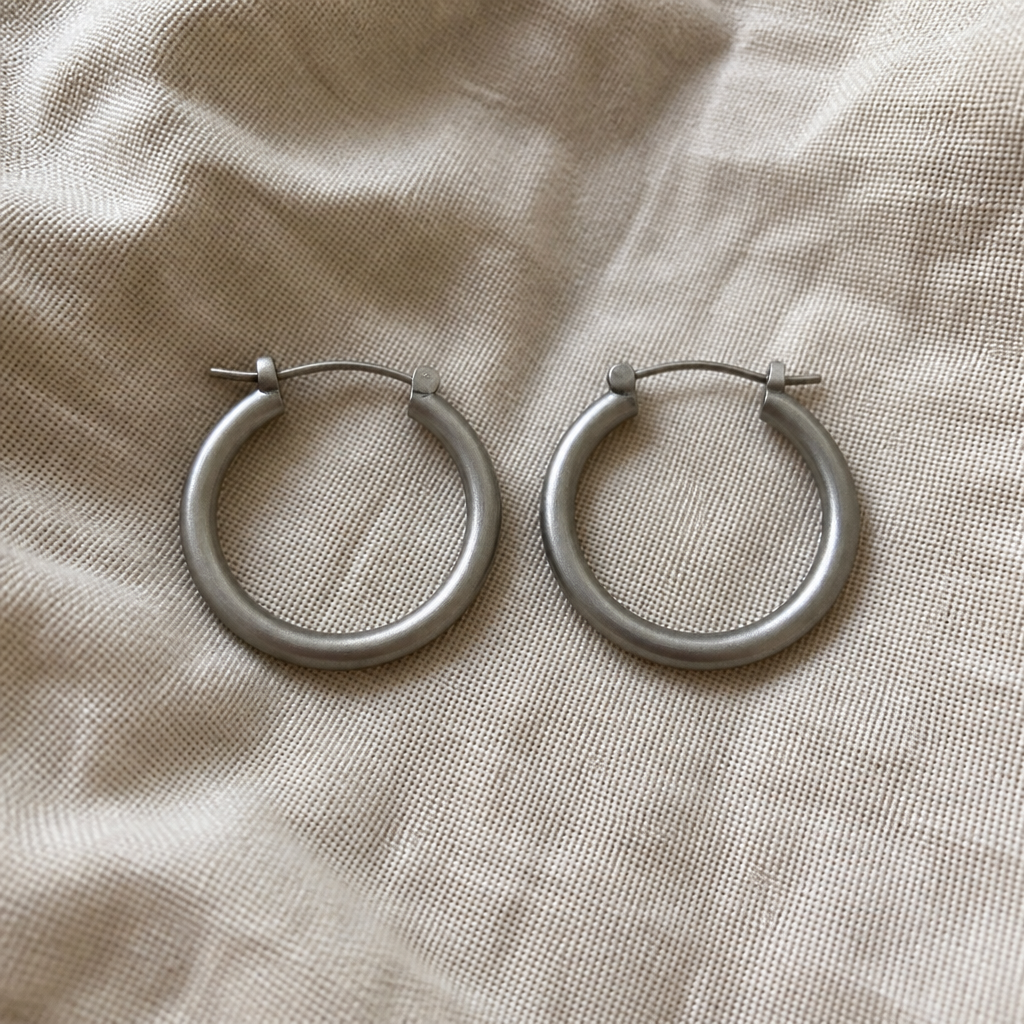 Side-by-side muted comparison of plain dull unbranded metal hoop earrings on a soft beige linen background showing no sparkle and no personality