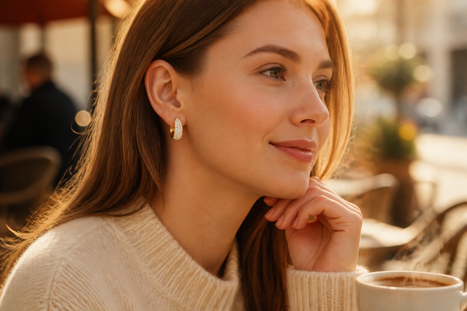 European woman in cream knit sweater at a sunlit cafe terrace wearing Dextera crystal pavé hoops, soft golden morning light on her face