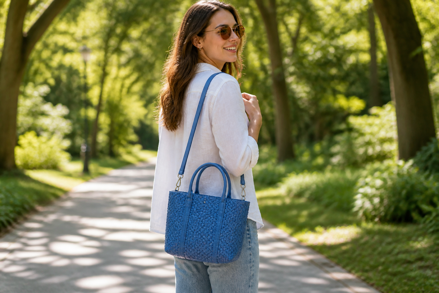 Model wearing the Mini Azure woven tote on her shoulder using the adjustable shoulder strap, walking through a sunny park with hands-free convenience and casual style