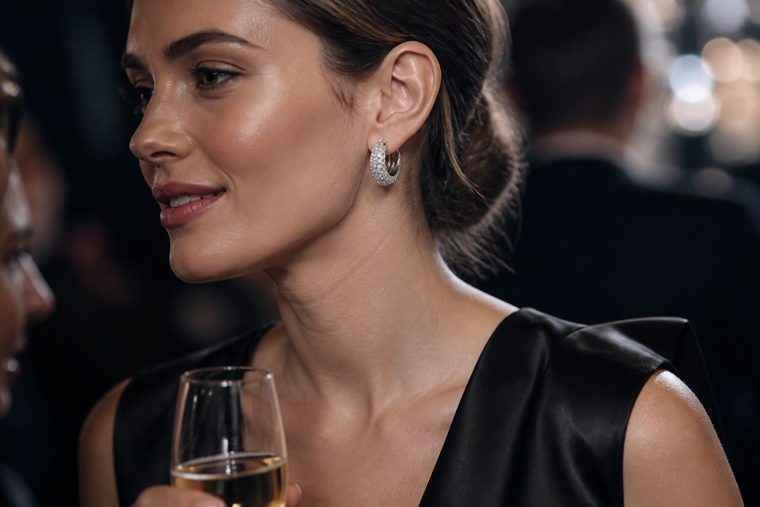 European woman in black silk dress at an evening event wearing Dextera pavé crystal hoops that catch and reflect ambient party lighting dramatically