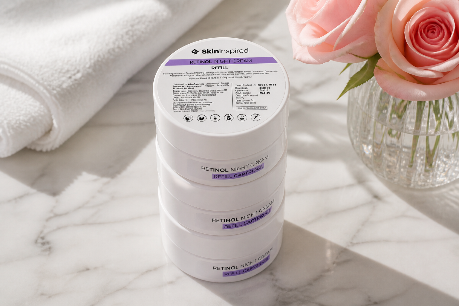 Lifestyle overhead photo of three stacked white retinol jars on a clean marble vanity with soft pink roses and a folded towel