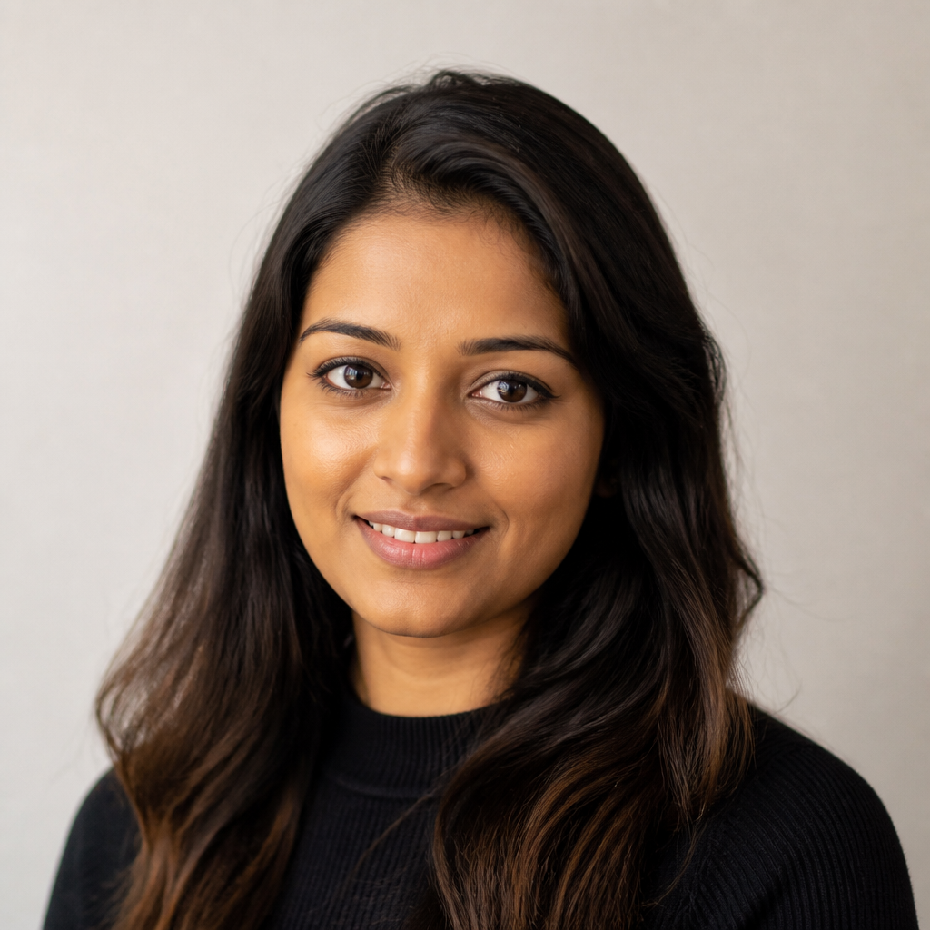 Headshot of a smiling young Indian woman with long dark hair, soft window light, neutral expression turning into a gentle laugh, warm cream backdrop
