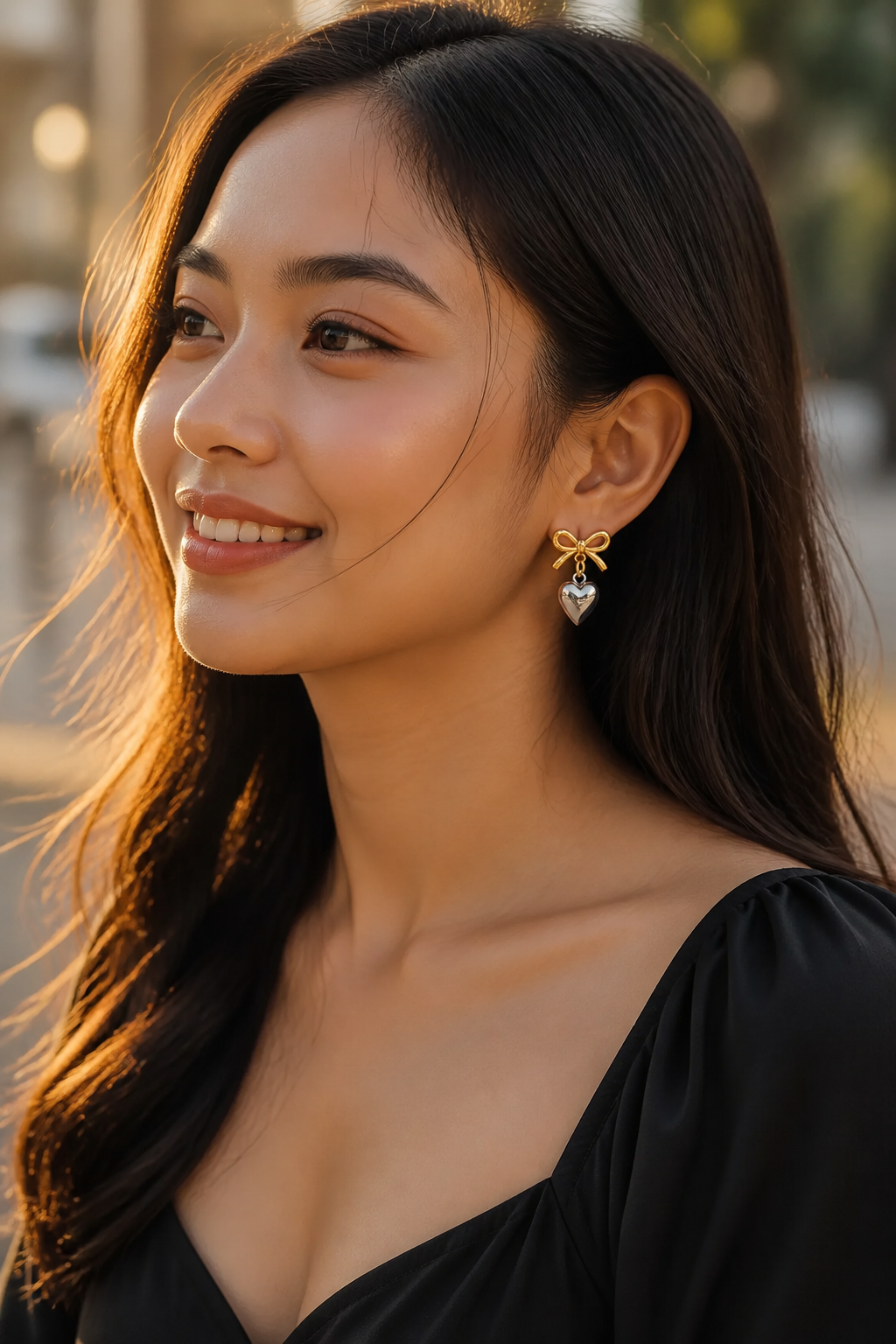 Southeast Asian woman smiling outdoors in a black dress wearing heart earrings, photographed in soft golden-hour light