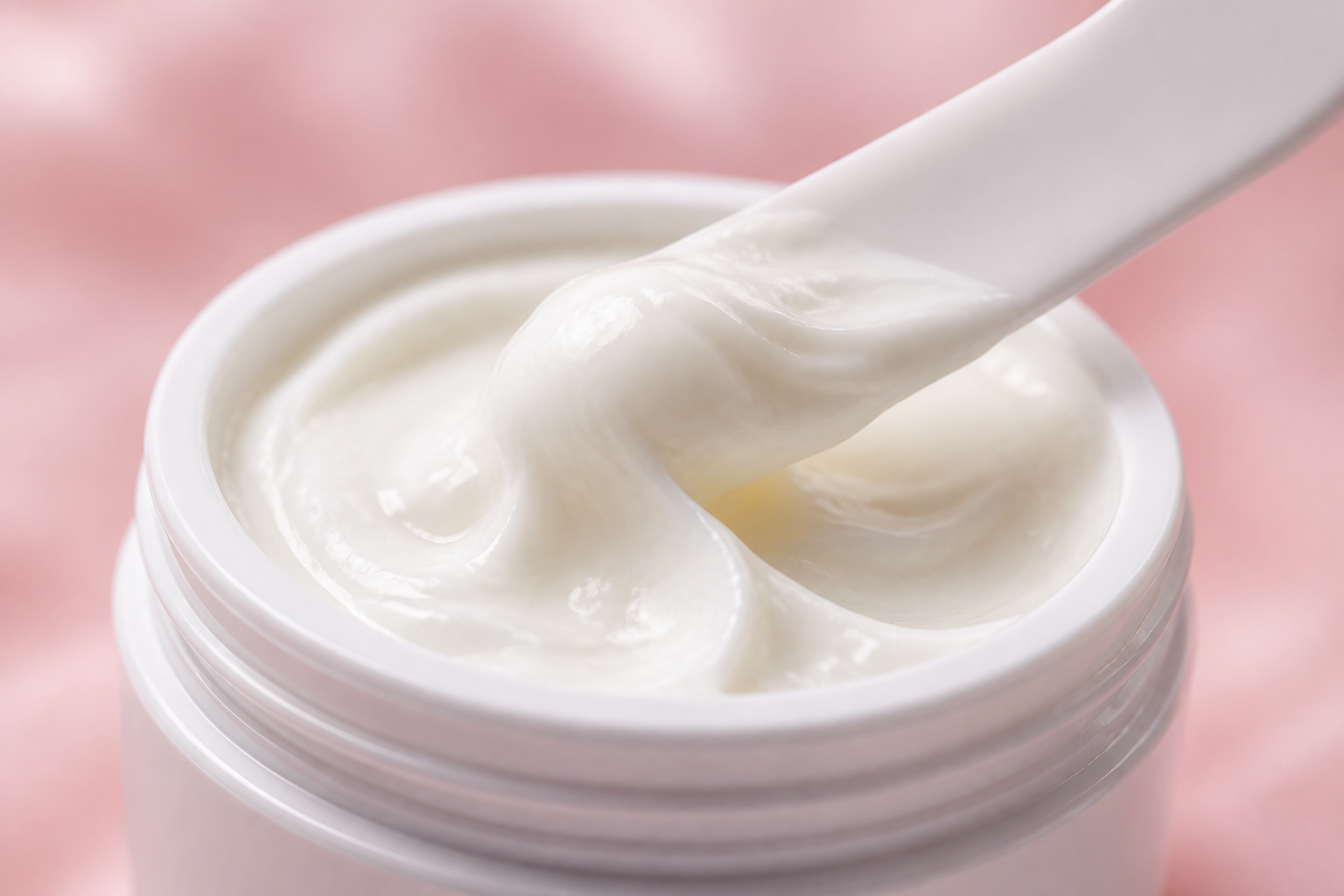Extreme close-up beauty shot of creamy white retinol cream being scooped from a luxe 50g glass jar with a soft pink backdrop