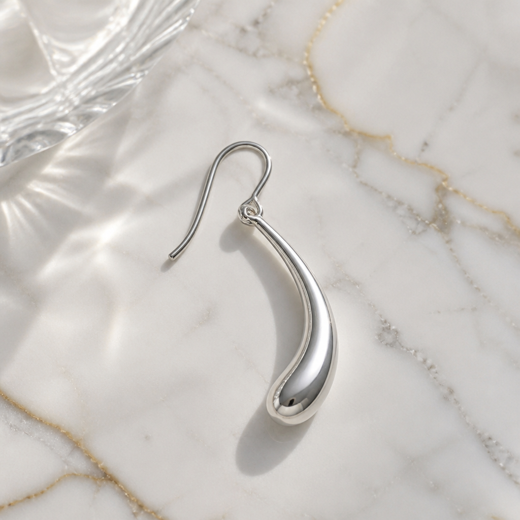 Silver Lumen Hook earring displayed on a polished marble surface with bright daylight reflecting off the curved metal finish