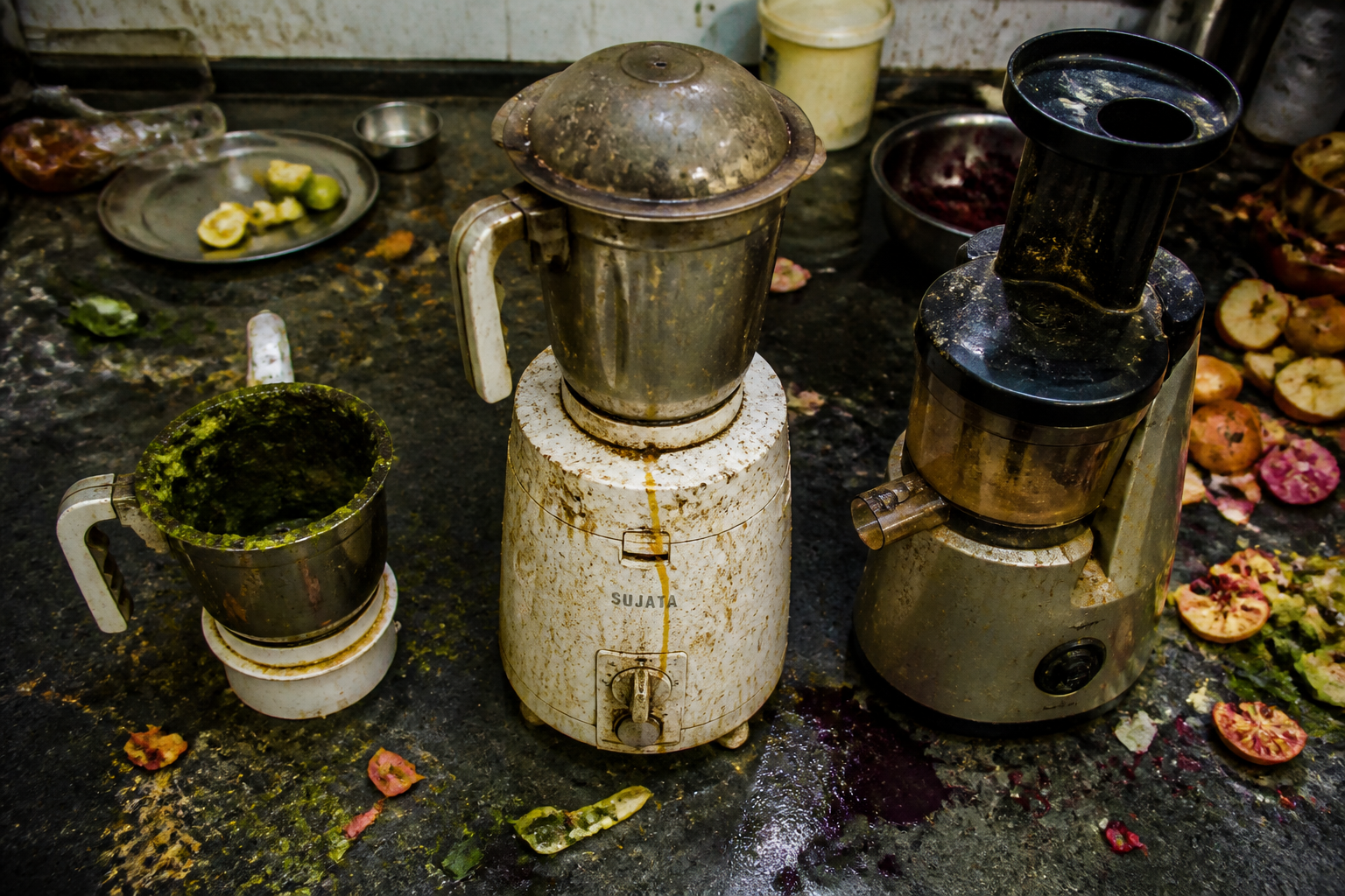 Wide overhead shot of a chaotic kitchen counter with an old rusty leaking mixer, a stained grinder with dried chutney on its rim, and a dented slow juicer surrounded by pulp splatter and fruit peels in unflattering overhead lighting